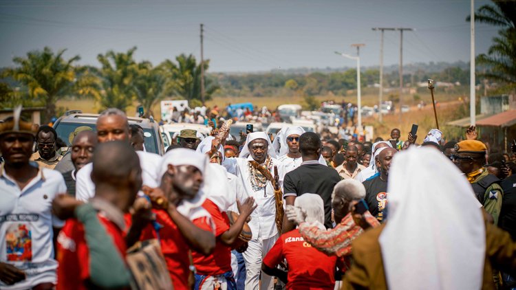 Mwata Kazembe steps out in bright white attire, layered beadwork, and ivory neck pieces catching the light, his presence announced before a word is spoken. (Image by Chona Mweemba for Nkwazi Magazine).